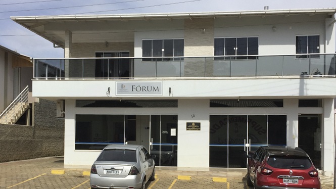 Foto horizontal: fachada do Fórum da Comarca de Campo Belo do Sul, com dois veículos, um prata e outro vermelho, estacionados em frente ao prédio.
