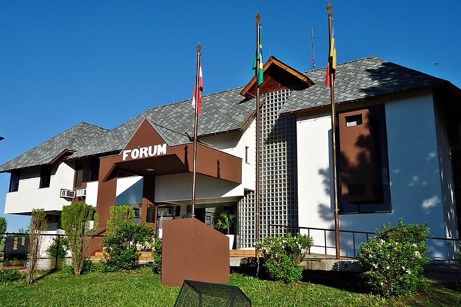 Foto horizontal: fachada do Fórum da comarca de Campo Erê, com entrada principal, jardins e mastros com bandeiras.