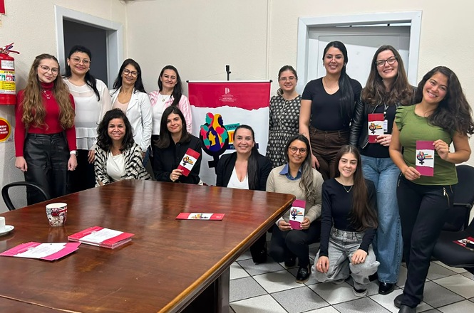 Foto horizontal: grupo de mulheres posa para registro coletivo, algumas segurando cartilhas, ao redor de mesa em sala de reunião.