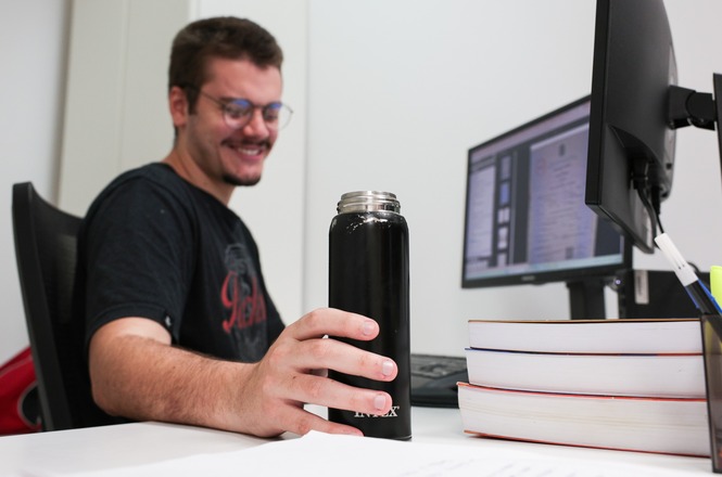  Copilot said: Horizontal: Pessoa utiliza garrafa reutilizável em estação de trabalho, ao lado de monitor, teclado e livros empilhados sobre a mesa. O ambiente é um escritório corporativo com mobiliário moderno, divisórias claras e equipamentos de informática voltados às atividades administrativas.