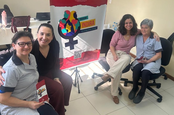 Foto horizontal: quatro mulheres posam juntas ao lado de banner do Programa Indira, duas delas abraçadas durante a atividade.