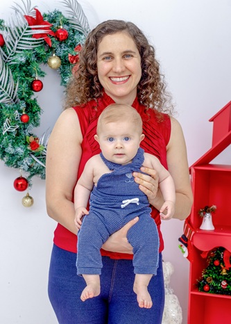 Foto vertical: mulher sorri enquanto segura bebê no colo diante de decoração natalina