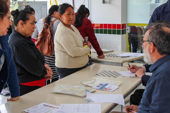  Copilot said: Foto colorida: pessoas em fila diante de mesa de atendimento, sala comunitária interna, ação social, aguardando orientação em branco, vermelho e verde