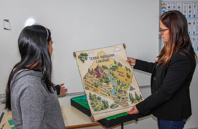 Foto colorida: pessoas observando mapa ilustrado de terra indígena, sala de aula interna, atividade educativa, apresentando território em verde, bege e marrom