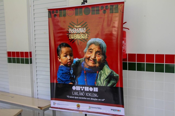 Foto horizontal: banner vermelho do projeto Cidadania Originária exibe mulher indígena com criança no colo.