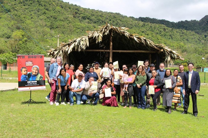 Foto horizontal: grupo posa com certificados diante de cabana de palha, cercada por gramado e morros verdes.