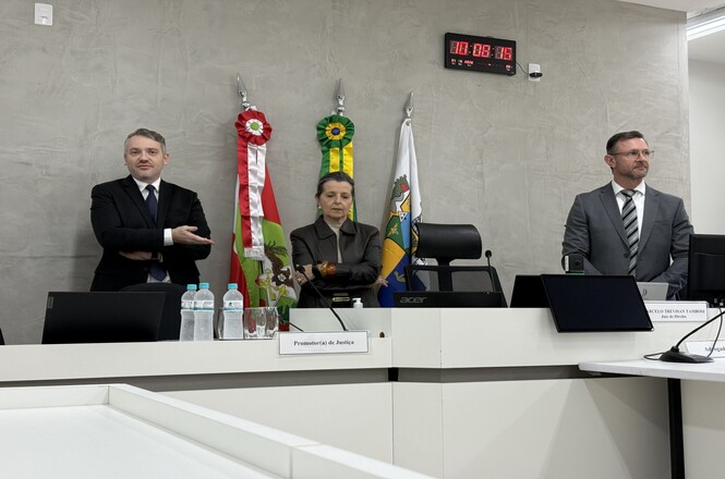 Foto horizontal: autoridades participam de ato institucional durante inspeção, mesa diretiva ao centro com bandeiras ao fundo, ambiente formal, clima de seriedade e cooperação.