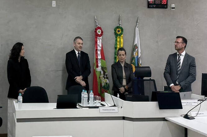 Foto horizontal: autoridades e servidores participam de ato institucional durante inspeção, alinhados atrás da mesa com bandeiras ao fundo, ambiente formal, clima de seriedade e cooperação.