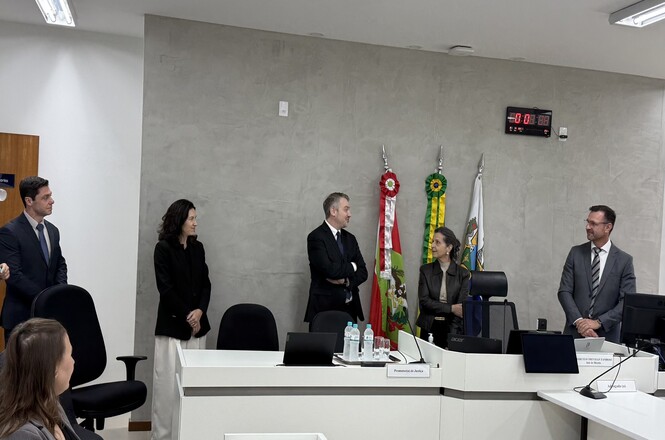 Foto horizontal: autoridades e servidores conversam durante ato institucional em inspeção, alinhados ao redor da mesa com bandeiras ao fundo, ambiente formal, clima de diálogo e cooperação.