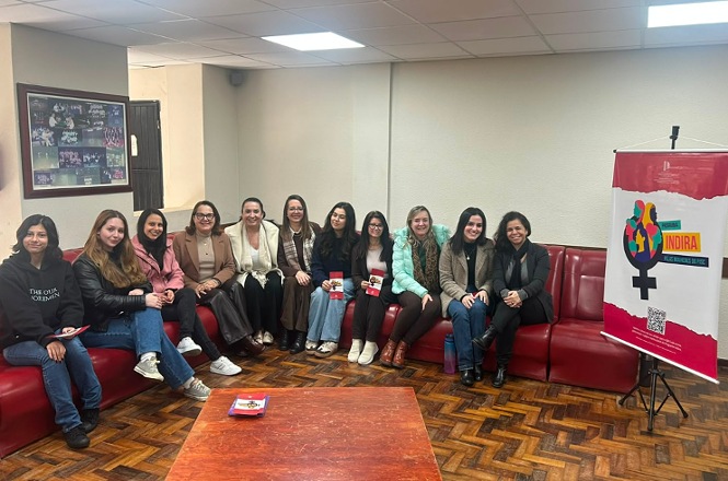 Foto horizontal: grupo de mulheres posa sentado em sofá, ao lado de banner do Programa Indira, durante atividade coletiva.