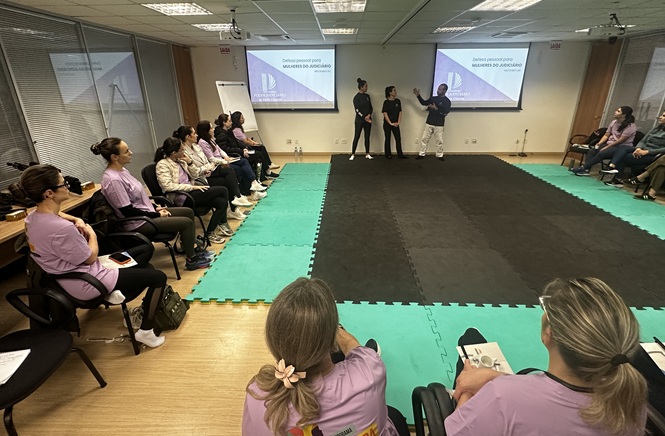Foto horizontal: grupo de mulheres acompanha orientações de três instrutores durante curso de defesa pessoal, em sala preparada com tatames.