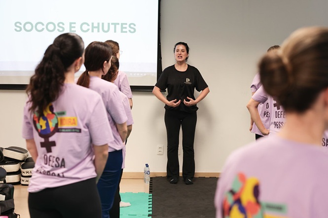 Foto horizontal: instrutora orienta grupo de mulheres durante atividade prática de defesa pessoal, com projeção sobre socos e chutes ao fundo.