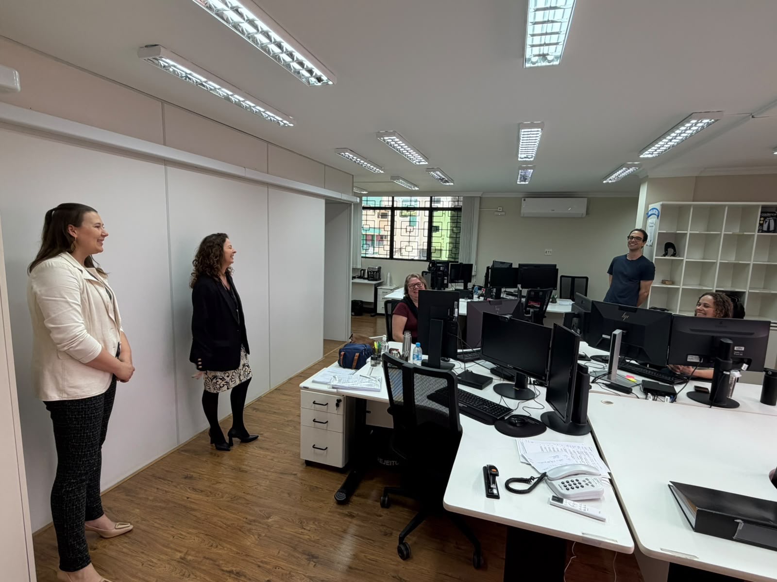 Foto horizontal: servidoras conversam com a equipe durante visita a setor administrativo, com mesas e computadores.