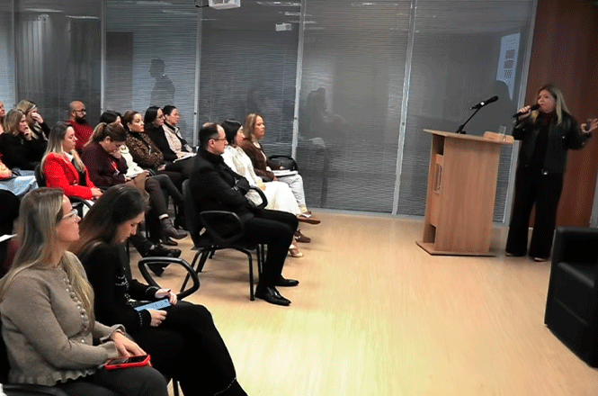 Foto horizontal: palestrante apresenta tema a público sentado em auditório, durante evento de capacitação em ambiente corporativo.