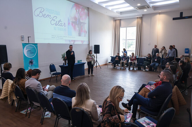 Foto horizontal: encontro do programa Bem-Estar no Trabalho, participantes em círculo e facilitadores ao centro, ambiente amplo, clima de integração.