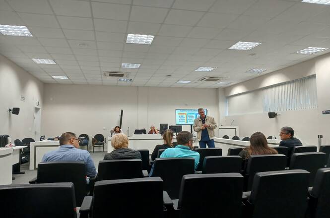 Foto horizontal: palestrante fala ao microfone diante de participantes reunidos em auditório, com apresentação projetada em tela ao fundo.