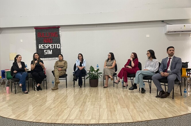 Imagem horizontal mostra autoridades em roda de conversa sobre bullying e respeito, diante de público escolar.