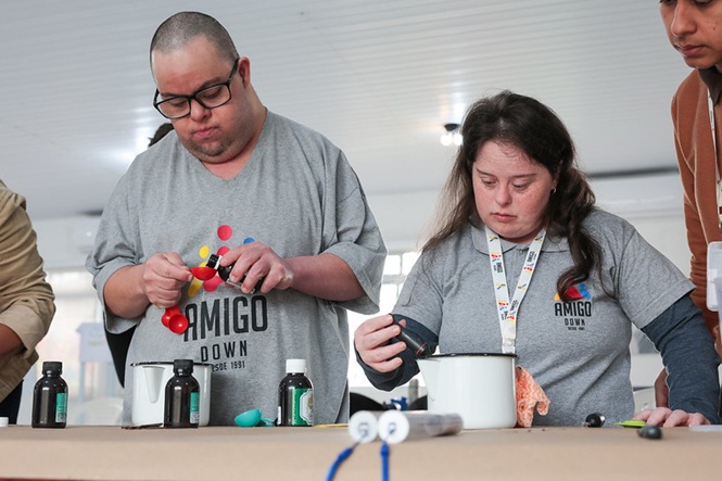 Foto horizontal: jovens com síndrome de Down participam de atividade prática, manuseando materiais durante oficina.