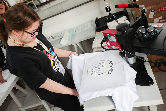 Foto horizontal: jovem com síndrome de Down segura camiseta personalizada ao lado de equipamento de estamparia durante oficina.