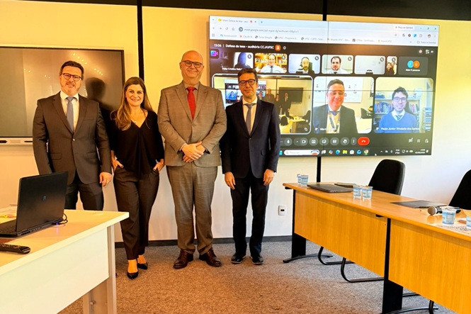 Foto horizontal: quatro integrantes de banca posam após apresentação acadêmica, com participantes da videoconferência exibidos em telão ao fundo.