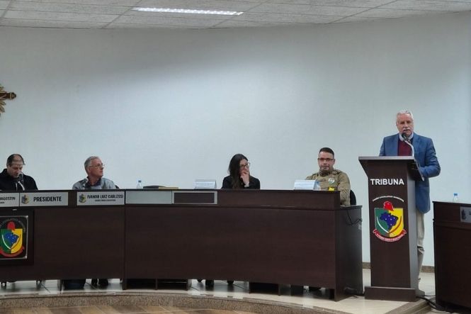 Foto horizontal: homem de terno azul fala ao microfone na tribuna da Câmara de Vereadores, diante de integrantes que compõem a mesa.