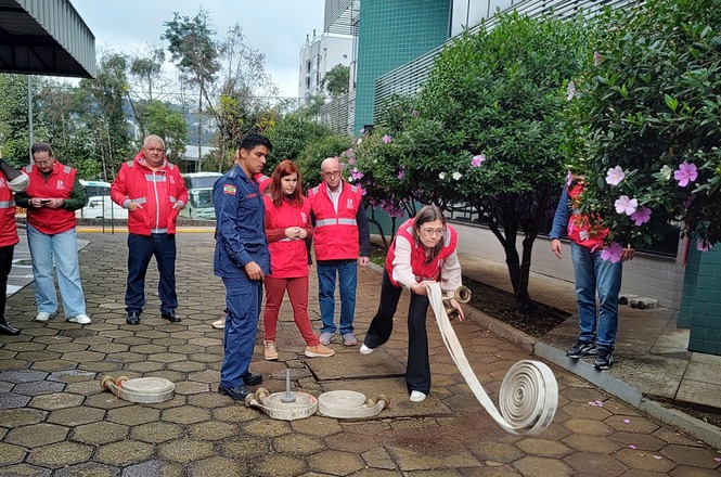 Foto horizontal: participante desenrola mangueira de incêndio durante treinamento prático, acompanhada por instrutor e colegas em atividade de prevenção.