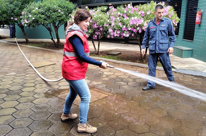 Foto horizontal: participante utiliza mangueira de incêndio durante treinamento prático, orientada por instrutor em área externa.