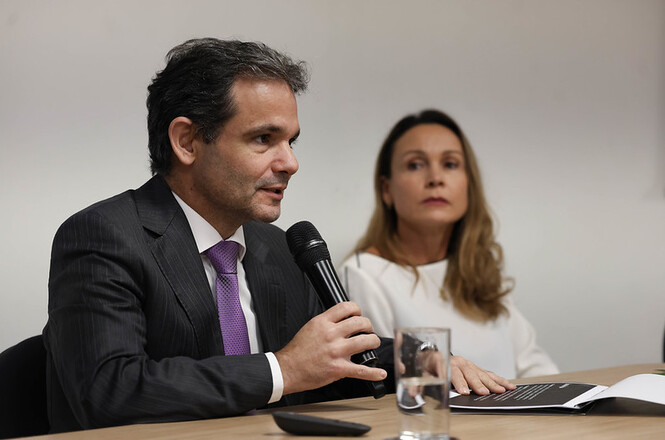 Foto horizontal: Des. Raphael Mendes Barbosa fala ao microfone durante mesa de debates, com participante sentada ao fundo.