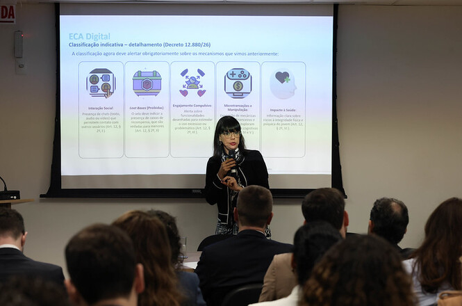 Foto horizontal: Mulher apresenta conteúdo em frente à tela de projeção enquanto participantes acompanham a palestra sentados no auditório.