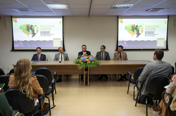 Foto horizontal: Cinco homens compõem mesa de autoridades em auditório, diante de público e telões com apresentação ao fundo.