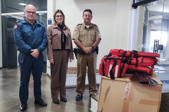 Foto horizontal: três agentes posam ao lado de kits de emergência sobre caixas, ambiente interno, tons neutros.