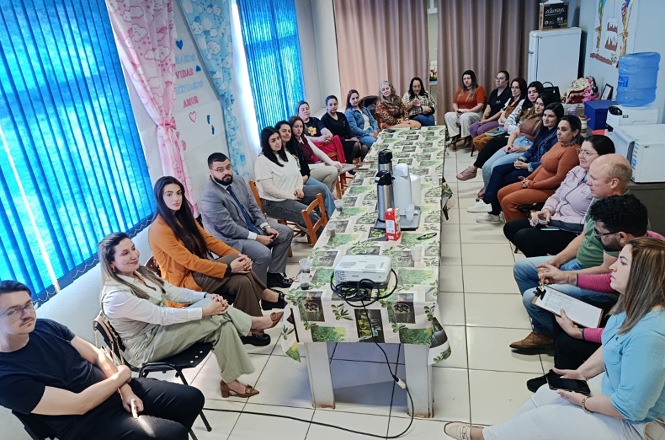 Foto horizontal: grupo participa de reunião, sentado ao redor de uma mesa, com projetor e materiais dispostos no centro.