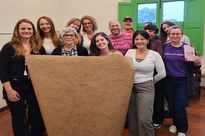 Foto horizontal: grupo posa em sala com cartaz de papel pardo preenchido por desenhos e palavras, diante de janelas verdes.