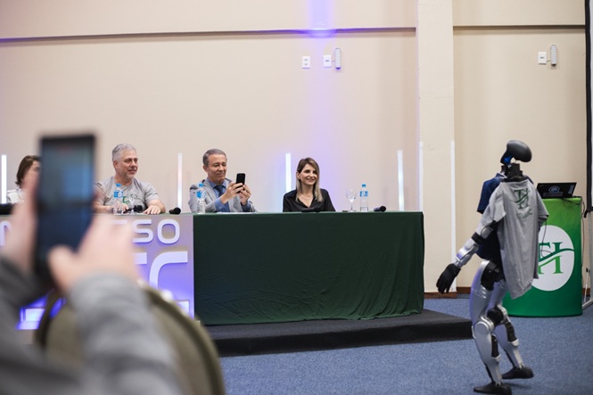 Foto horizontal: robô interage com participantes durante o Congresso Atjusc, enquanto integrantes da mesa acompanham a apresentação.