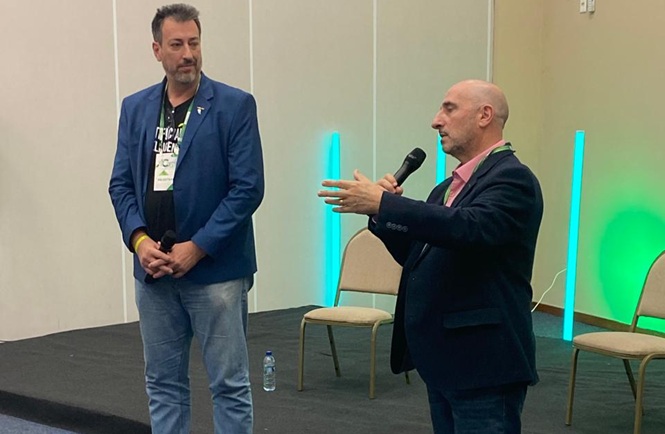 Foto horizontal: dois homens participam de apresentação no Congresso Atjusc, um deles fala ao microfone enquanto o outro acompanha.
