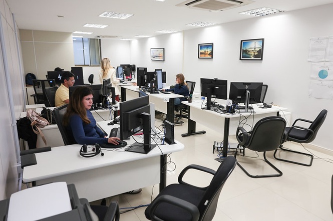 Foto horizontal: servidores trabalham em sala de atendimento da Vara Estadual de Execuções Fiscais Municipais, com mesas brancas, computadores, cadeiras pretas e quadros nas paredes.