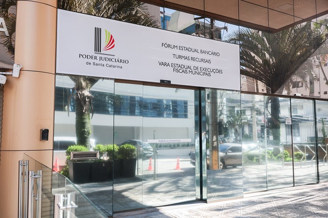 Foto horizontal: fachada envidraçada do Fórum Estadual Bancário, Turmas Recursais e Vara Estadual de Execuções Fiscais Municipais, com identificação do Poder Judiciário de Santa Catarina acima da entrada principal.