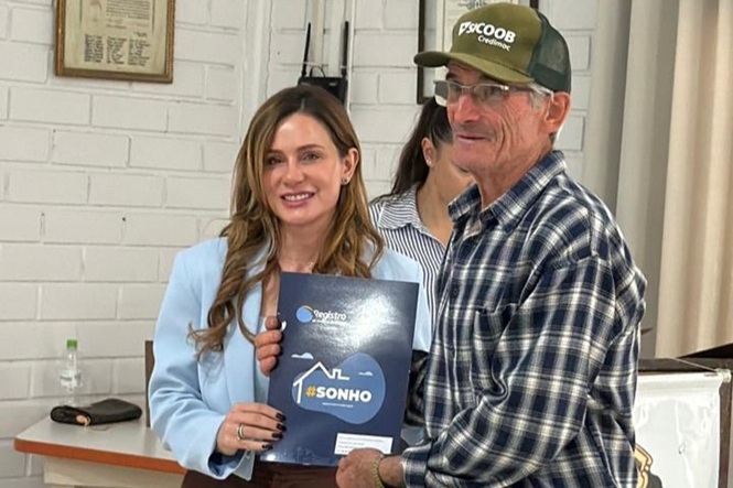 Foto horizontal: mulher de blazer azul-claro e homem de camisa xadrez e boné posam sorridentes, segurando cartilha do programa Lar Legal com a inscrição “#SONHO”.
