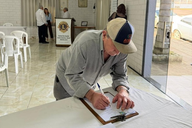 Foto horizontal: homem de boné inclina-se sobre mesa enquanto assina documento preso a uma prancheta, em salão com cadeiras e pessoas ao fundo.
