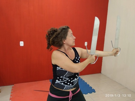 Mulher pratica treino com espadas em ambiente interno, postura firme e foco, clima de disciplina e atividade física