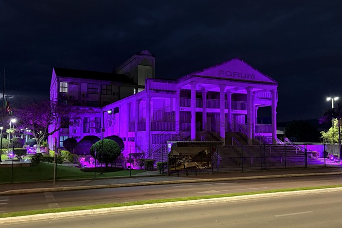 Foto horizontal noturna: fachada do Fórum de Criciúma iluminada em tons de roxo durante ação de conscientização.