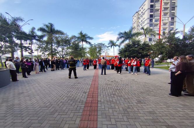 Foto horizontal: participantes reunidos em área externa durante simulado de evacuação, equipes organizadas, clima de prevenção e segurança coletiva.