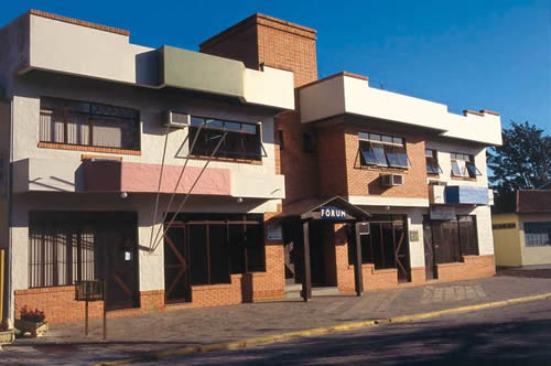 Foto horizontal: fachada do Fórum da Comarca de Garopaba, em tons de branco, rosa e tijolos aparentes, com janelas e aparelhos de ar-condicionado.
