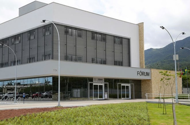 Foto horizontal: fachada do fórum de Garuva, prédio institucional moderno com área verde ao redor, clima de acesso à Justiça.