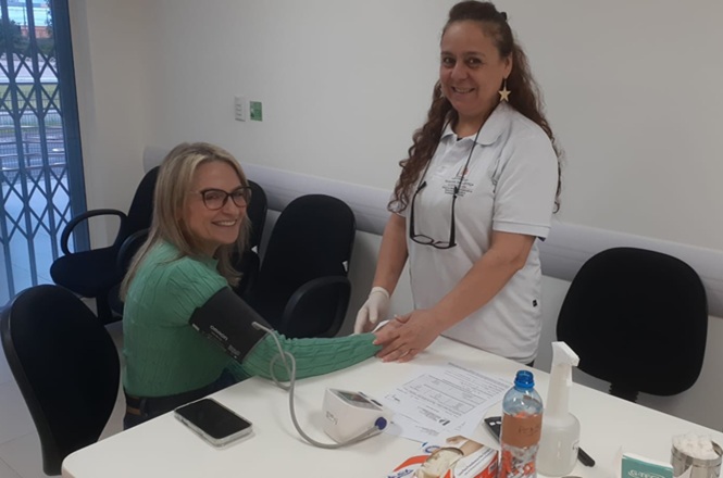 Foto horizontal: profissional de saúde em camisa polo branca e luvas afere a pressão arterial de servidora em blusa verde sentada à mesa em sala clara com cadeiras pretas durante ação de atendimento institucional.