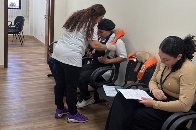 Foto horizontal: profissional de saúde em camiseta branca e calça preta ajusta aparelho massageador laranja nos ombros de funcionária com touca preta sentada em cadeira preta em sala clara de piso amadeirado durante atividade institucional de bem-estar.