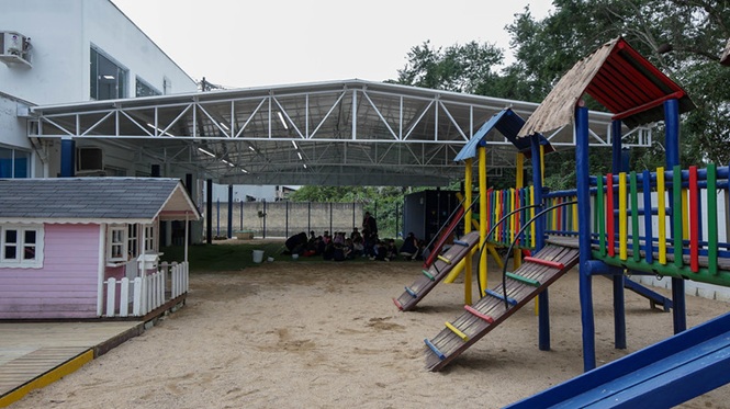 Foto colorida: playground com escorregadores coloridos e casinha infantil, pátio escolar coberto, recreação educativa, oferecendo espaço de atividades em azul, amarelo e rosa.