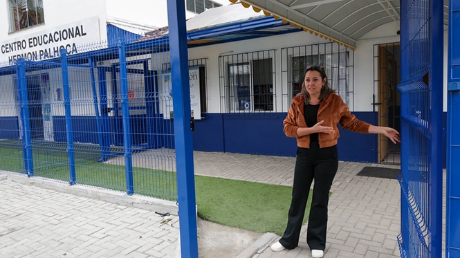 usando casaco marrom, blusa preta e calça escura em frente ao Centro Educacional Hermon Palhoça, área externa escolar, apresentando a entrada em azul, branco e marrom.