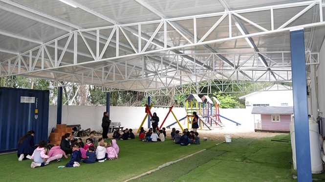 Foto colorida: crianças sentadas em grupos sob cobertura escolar, pátio com playground ao fundo, atividade educativa, participando de dinâmica em azul, verde e rosa