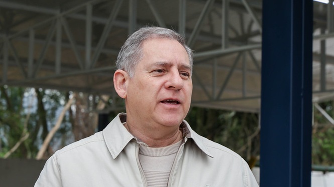 Foto colorida: homem com cabelos curtos grisalhos, usando jaqueta clara sobre blusa de tom bege, em área externa coberta de instituição educacional, falando diante da câmera.
